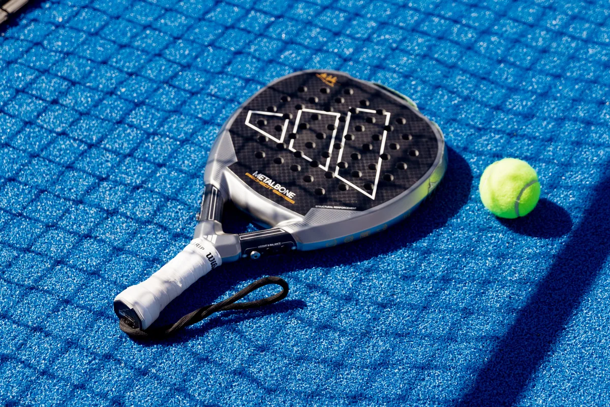 A yellow ball beside a perforated padel racket on a blue court